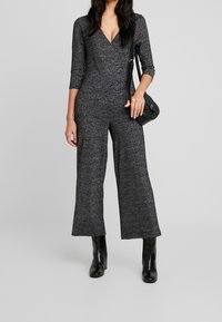 Mono gris ajustado con escote en V profundo, mangas tres cuartos y piernas anchas. Combinado con botas de tacón negras y un bolso pequeño.