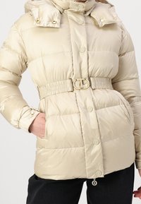 Beige péřová bunda s odnímatelnou kapucí, lesklým povrchem, elastickým pasem se zlatými detaily a knoflíkovým zapínáním. Obsahuje boční kapsy.