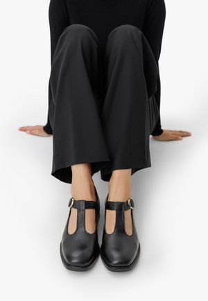 Person sitzt mit gebeugten Beinen, trägt schwarze weit geschnittene Hosen und schwarze T-Riemen-Schnallenschuhe auf einer weißen Fläche.