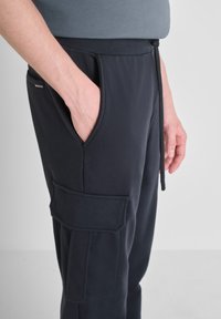 Mornarske hlače za trčanje s elastičnim pojasom, vrpcom, bočnim džepovima i velikim cargo džepom na lijevoj nozi, izrađene od mekanog materijala.