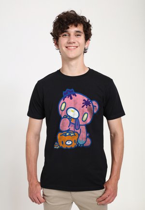 Ung mand i en sort t-shirt med en farverig grafik af en lyserød tegneserie-bjørn, der holder en orange genstand, smilende og stående mod en ensfarvet baggrund.