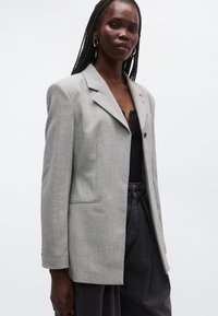 Γκρι σακάκι με δομημένους ώμους, κλείσιμο με ένα κουμπί και υφασμάτινη υφή. Διαθέτει γιακά με σκίσιμο και πλαϊνές τσέπες.
