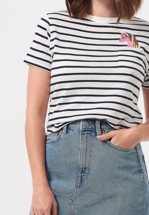 Femme portant un t-shirt à rayures blanches et bleu marine avec une licorne colorée brodée près de la poche poitrine, associé à une jupe en denim taille haute.