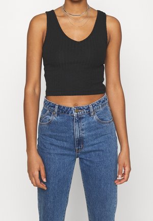 Vrouw draagt een zwart geribbeld cropped tanktopje, gelaagde zilveren kettingen en blauwe high waist spijkerbroek met knoopsluiting.