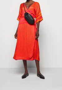 En knälång klänning i klar orange med mönstrad textur, med vida ärmar och en distinkt v-ringning. Svart axelremsväska och svarta lägenheter.