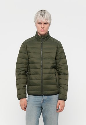 Fiatal férfi, rövid platinaszőke hajjal, zöld steppelt cipzáros dzsekit és világoskék farmert visel, egyszínű szürke háttér előtt áll.