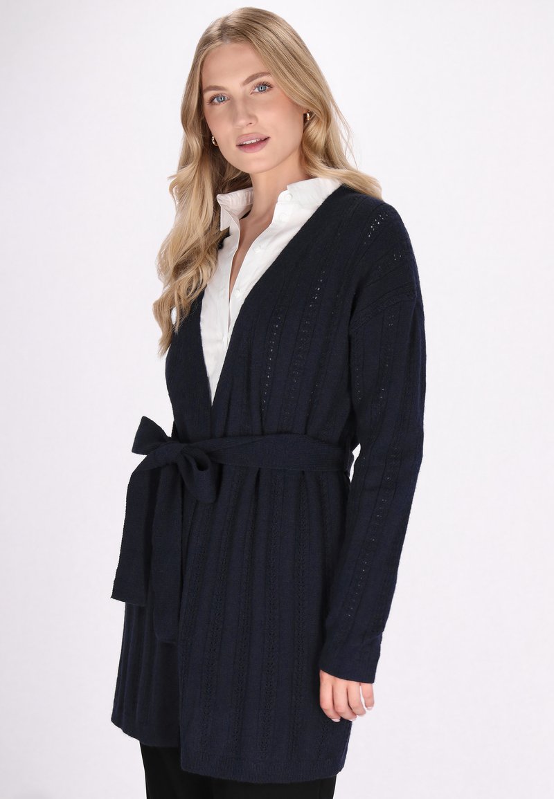 Cardigan lavorato a maglia di colore blu navy con vita cintata, apertura frontale e trama a coste verticali. Presenta maniche lunghe con dettagli intricati.