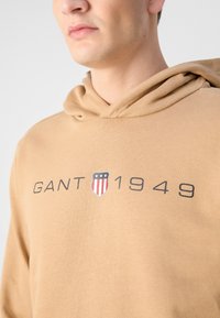 Felpa beige con un logo caratterizzato da strisce rosse e blu e il testo "GANT 1949" in grigio, realizzata in un tessuto morbido con una vestibilità comoda.