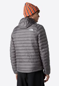 Grå vadderad jacka med horisontell quiltad design, huva och logotyp på den övre delen av ryggen, tillsammans med en flerfärgad randig stickad mössa.