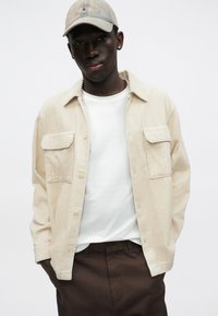 Bézs kordbársony ing két elülső zsebbel, fehér póló alatta, és barna nadrág; kiegészítve világos baseball sapkával.