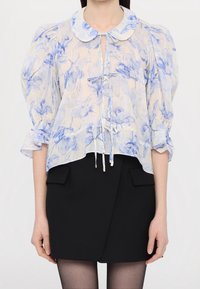 Lekka bluzka z niebieskimi kwiatowymi wzorami na kremowym tle, posiada szpiczasty kołnierzyk oraz wiązanie przy dekolcie. Bufiaste rękawy.