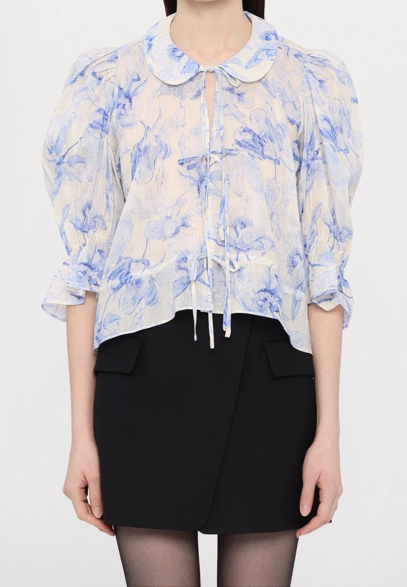 Lekka bluzka z niebieskimi kwiatowymi wzorami na kremowym tle, posiada szpiczasty kołnierzyk oraz wiązanie przy dekolcie. Bufiaste rękawy.