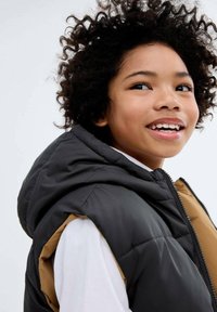 Vest puf negru și căprui cu glugă, având un fermoar frontal și o textură moale. Purtați peste o cămașă albă.