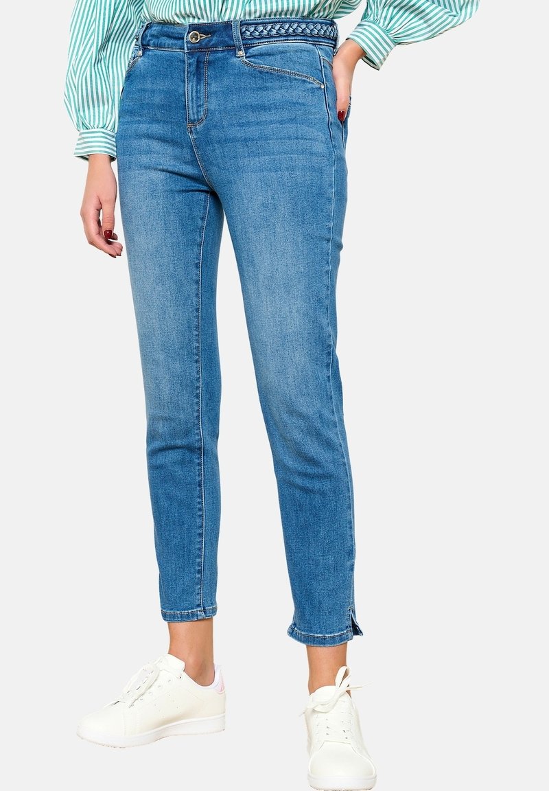 Vaaleansiniset farkut, joissa on istuva malli, keski-vyötärö, punottu vyö ja nilkkapituiset lahkeet, joissa on halkiot. Yhdistettyinä valkoisiin lenkkareihin.