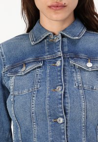 Giacca in denim di colore blu chiaro, colletto classico, chiusura frontale con bottoni, due tasche sul petto e dettagli di cucitura visibili.
