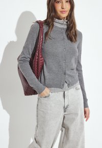 Grijze gebreide cardigan met knoopsluiting, gedragen over een licht shirt. Gecombineerd met lichtgrijze wijde jeans en een gestructureerde bordeauxrode tote bag.