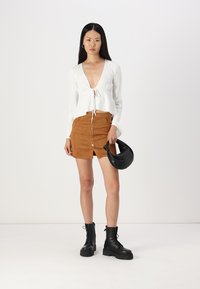 Haut à manches longues blanc côtelé avec un détail de nœud, assorti à une mini jupe en daim marron avec un devant à boutons. Sac à main rond noir et bottes de combat.