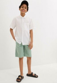 Camicia bianca a maniche corte, pantaloni corti verde menta, sandali neri e un cappellino nero. Sfondo bianco uniforme.