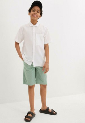 Chemise blanche à manches courtes, shorts vert menthe, sandales noires et casquette noire. Fond blanc uni.