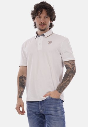 Uomo con capelli ricci che indossa una polo bianca e jeans blu, con tatuaggi visibili su entrambi gli avambracci, in piedi con una mano in tasca.