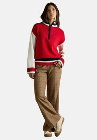 Roter Pullover mit einem halben Reißverschluss, weißen Ärmeln und marineblauen Akzenten. Kombiniert mit braunen karrierten Schlaghosen und cremefarbenen Sneakern.