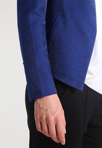 Blazer structuré bleu avec un tissu légèrement texturé, doté d'une poche avant unique et d'une coupe droite. Le col n'est pas visible.