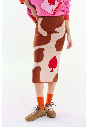 Dzianinowa midi spódnica z abstrakcyjnymi brązowo-beżowymi kształtami i czerwonym symbolem pika, zestawiona z pomarańczowymi skarpetkami, brązowymi tenisówkami i różowym swetrem.