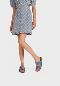 Sandali blu con doppie cinghie regolabili e una soletta testurizzata, abbinati a un vestito in denim con un design fantasia e maniche corte.