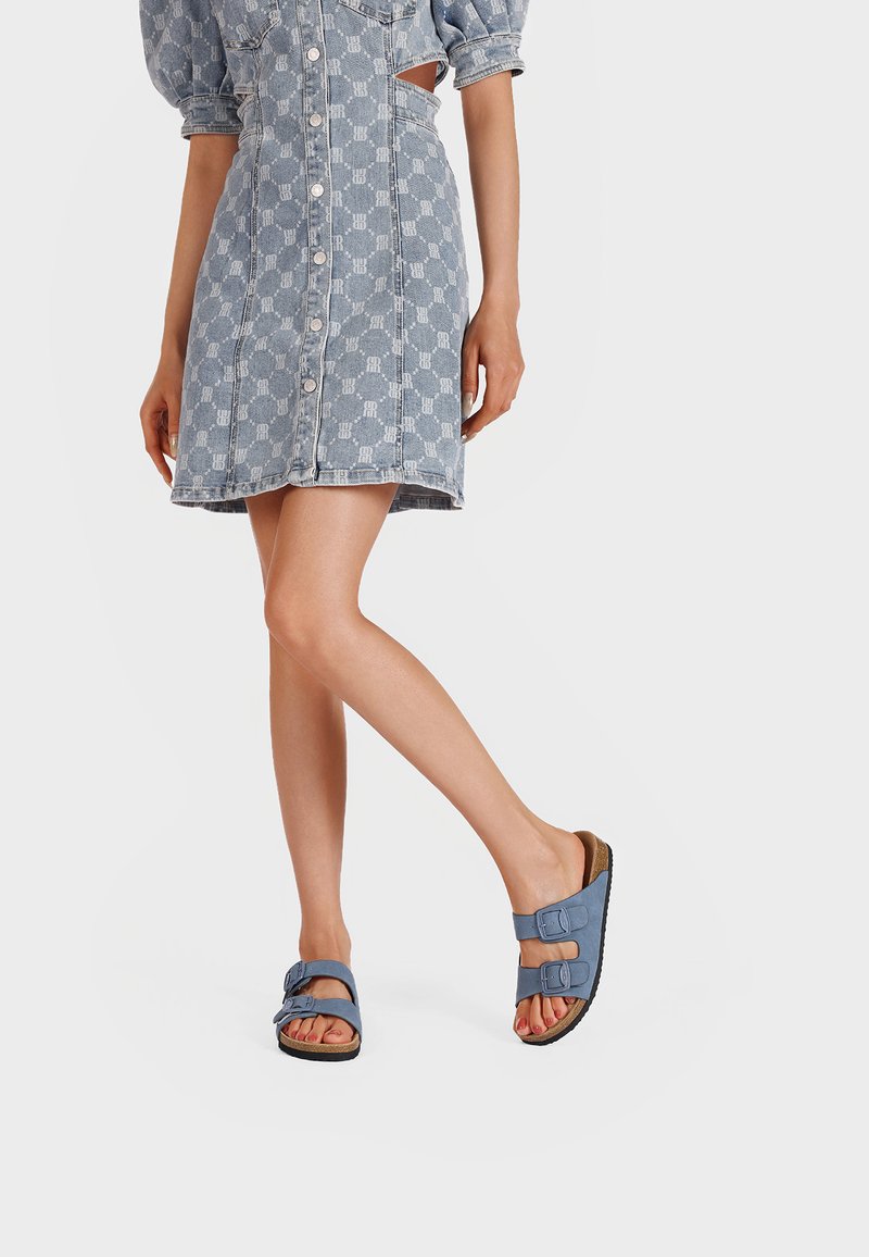 Sandali blu con doppie cinghie regolabili e una soletta testurizzata, abbinati a un vestito in denim con un design fantasia e maniche corte.