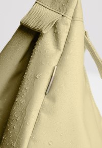 Borsa in tessuto beige con gocce d'acqua sulla sua superficie, evidenziando la sua texture impermeabile e il dettaglio della tracolla.