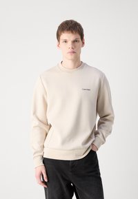 Béžový sveter s okrúhlym výstrihom, dlhými rukávmi a malým čiernym logom "Calvin Klein" na hrudi, skombinovaný s tmavými džínami.