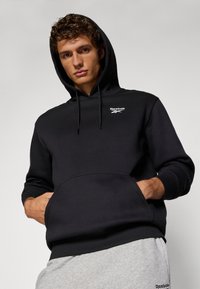 Čierna mikina s kapucňou a predným vreckom, s rebrovanými manžetami a lemom. Obsahuje malé biele logo Reebok na hrudi. Mäkká textúra látky.
