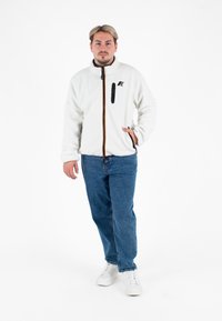 Biała polarowa kurtka z czarnym zamkiem, pomarańczowym i żółtym paskiem akcentującym oraz kieszenią na piersi, zestawiona z niebieskimi dżinsami i białymi trampkami.