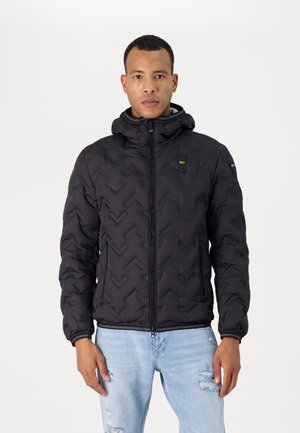 Giubbotto imbottito nero con texture a chevron, chiusura frontale con zip, cappuccio regolabile e tasche laterali, indossato sopra jeans chiari strappati.