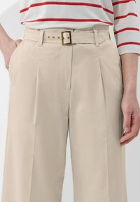 Beige Plissee-Hosen aus glattem Stoff, ausgestattet mit einem rechteckigen, goldfarbenen Schnallen-Gürtel und Seitentaschen.