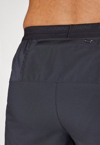 Acercamiento a la parte baja de la espalda de una persona que lleva puesto un pantalón corto deportivo negro con un bolsillo con cremallera y un panel lateral texturizado.