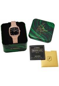 Montre Fossil avec un cadran carré noir, bracelet en maille rose doré ; emballée dans une boîte en métal marbré vert portant une étiquette noire et dorée, incluant une carte supplémentaire.