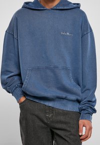 Felpa con cappuccio in tessuto blu sbiadito simile al denim, con tasca frontale e polsini a coste, decorata con un piccolo logo ricamato sul petto.