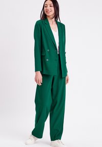 Green double-breasted blazer with notched lapels, paired with loose-fitting green trousers. Worn with a white v-neck shirt and white sneakers.