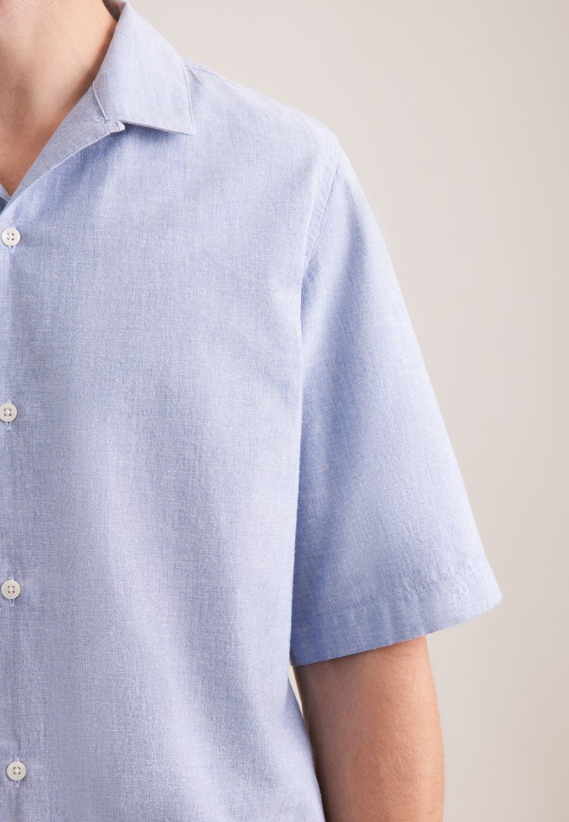 Chemise bleu clair à manches courtes avec boutons, portée par une personne, visible du cou jusqu'au milieu du torse sur un fond uni.