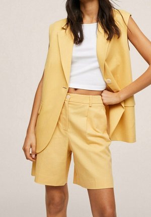 Vrouw die een gele mouwloze blazer en bijpassende shorts met een hoge taille draagt, met een wit cropped tanktop, staand met één hand in de zij.