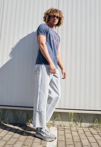T-shirt de sport bleu avec des accents et un logo orange, associé à un pantalon loose gris clair et des baskets grises. Texture lisse et coupe décontractée.