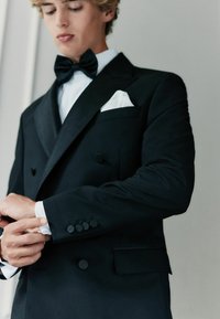 Schwarze Zweireiher-Tuxedojacke, strukturiertes Material, satinierte Revers, vier Knöpfe an den Manschetten, weißes Einstecktuch, getragen mit einer Fliege.