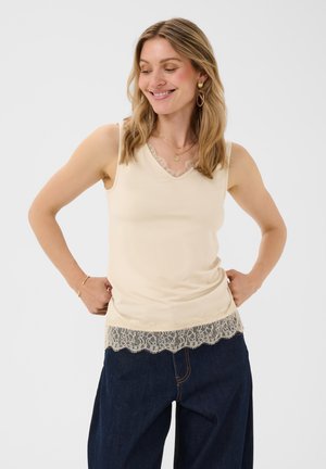 Femme souriante, portant un haut sans manches crème à bordure en dentelle et un jean bleu à jambes larges, les mains reposant sur les hanches, sur fond uni.