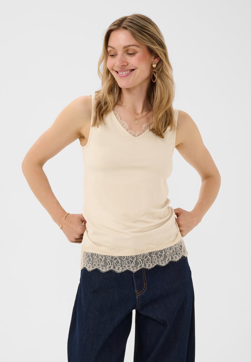 Femme souriante, portant un haut sans manches crème à bordure en dentelle et un jean bleu à jambes larges, les mains reposant sur les hanches, sur fond uni.