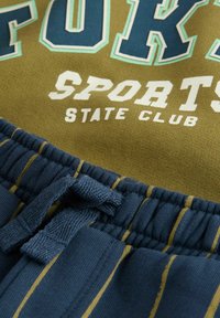 Zelený top s nápisem "SPORTS STATE CLUB" v bílé a modré barvě, spárovaný s námořnickými šortkami se svislými pruhy a detaily na šňůrce.