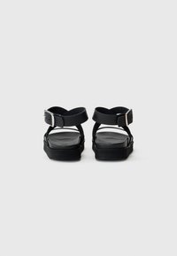 Sorte sandaler med to brede stropper og justerbare spenner. Flat såle med teksturert fotseng, minimalistisk design, og ingen distinkte mønstre.