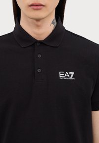 Čierna polokošela s gombíkovým golierom, s bielym logom EA7. Vyrobená z hladkej bavlnenej zmesi so skrátenými rukávmi a priliehavým strihom.