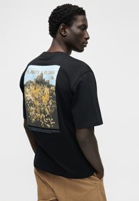 Μαύρο βαμβακερό t-shirt με γραφικό τοπίο λουλουδιών στην πλάτη, με μπλε και κίτρινες πινελιές, και ουδέτερο κείμενο κάτω από την εικόνα.