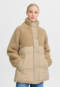 Beige puffjakke med en blød, fluffy tekstur på toppen, med en høj krave, trykknapper og to frontlommer.